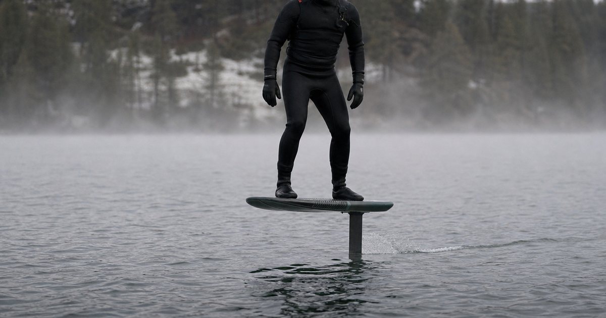 Rider in a thick black hooded wetsuit and gloves standing on a hydrofoil board above a gray-green lake, mist on the water, pine trees on the far shore, overcast winter light, no text