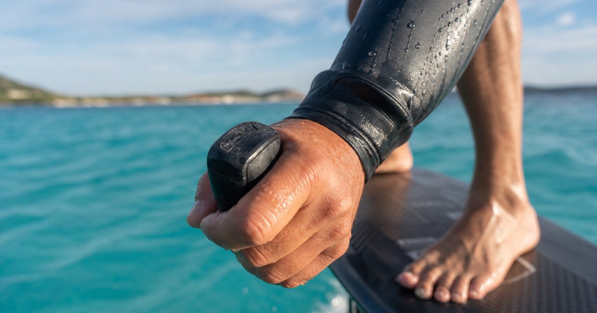 Close-up of a rider's wet hand gripping a compact wireless hand throttle above turquoise water, hydrofoil board visible behind, bright morning sunlight, no text