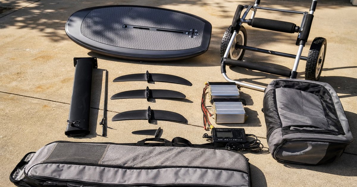 Complete hydrofoil kit laid out on a sunlit garage floor: board, mast, several front wings, two battery packs, charger, padded bags, and a folding beach cart, no text